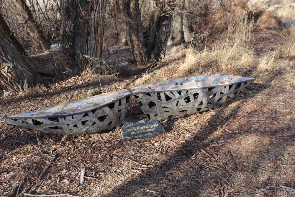 A metal sculpture, evocative of a canoe with trees in the background. There is a plaque that reads 