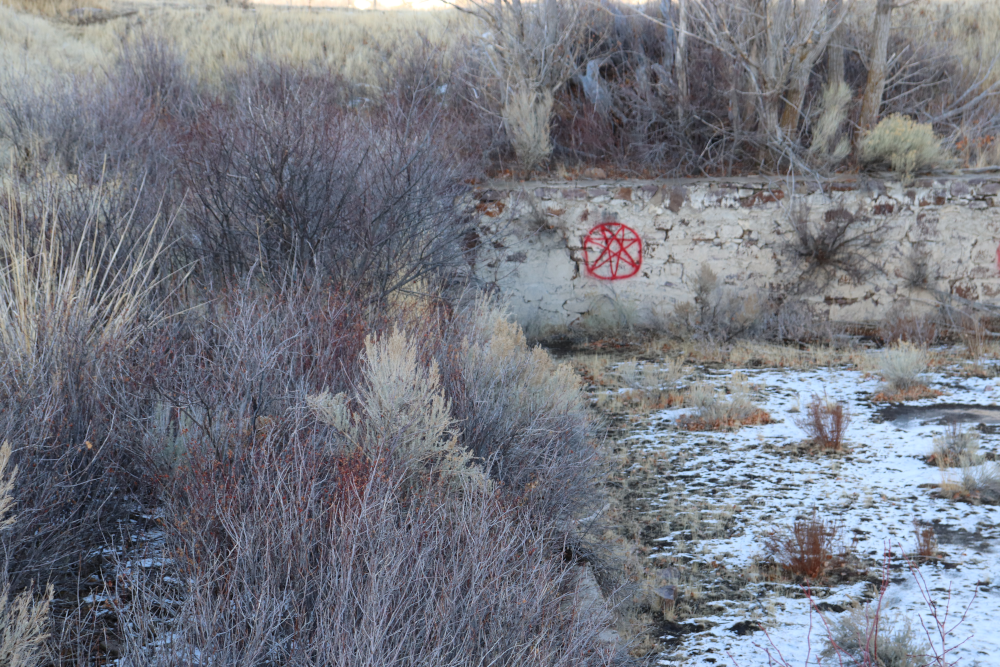A section of gray brick wall, surrounded by snow and sagebrush. Somebody has spray painted a star with seven points and a circle around it in red. 
