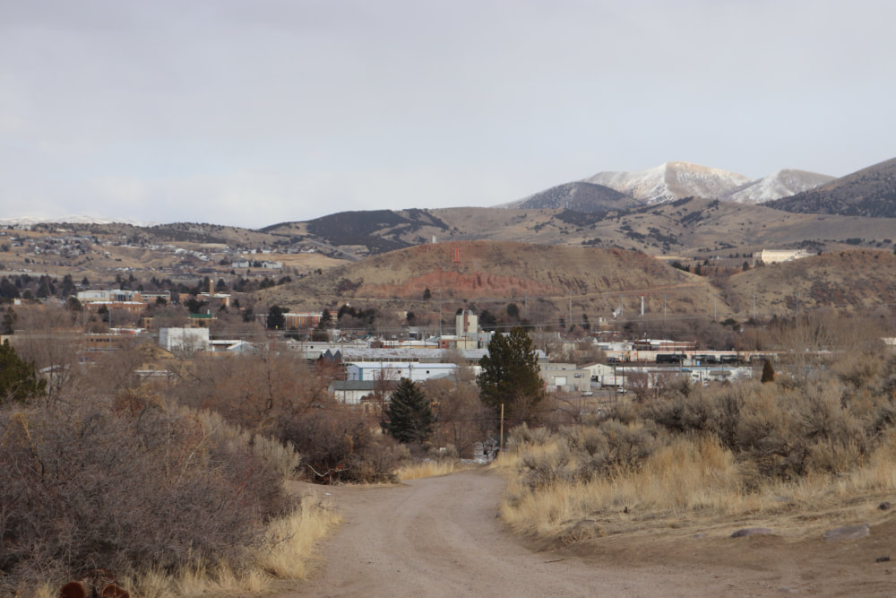 A dirt trail in the foreground, surrounded by sagebrush and scrubby plants leads to buildings. In the center, a red-tinted hill has an 